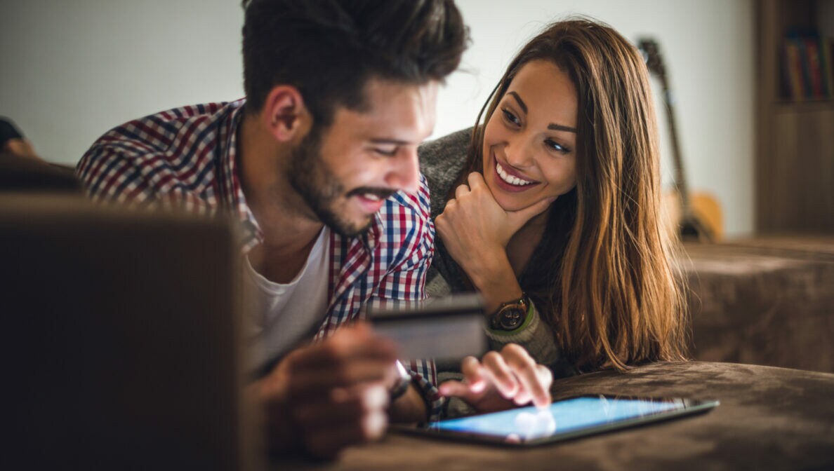 Ein Pärchen lächelt sich über ein Tablet an. Der Mann hält eine Kreditkarte.
