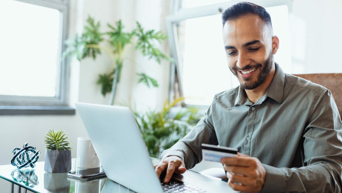 Eine Person sitzt an einem Schreibtisch vor einem Laptop, hält eine Kreditkarte in der Hand und lächelt.