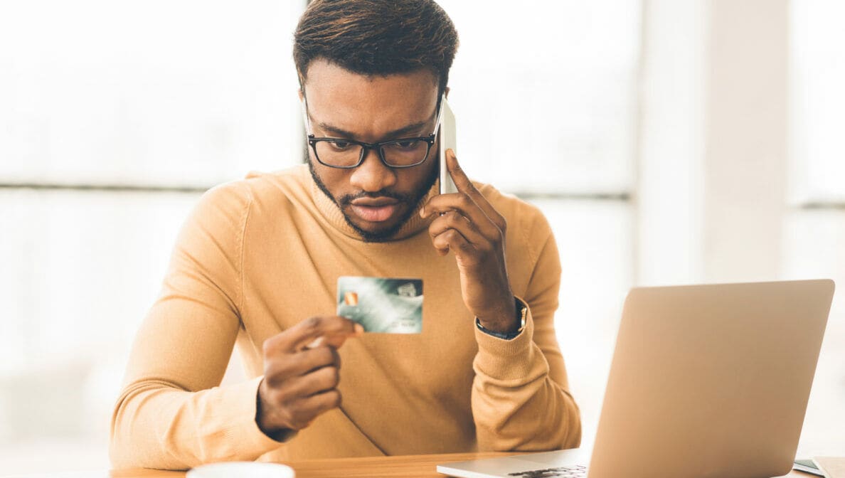 Eine Person sitzt telefonierend vor einem geöffneten Laptop am Tisch und blickt auf seine Bankkarte in der Hand.