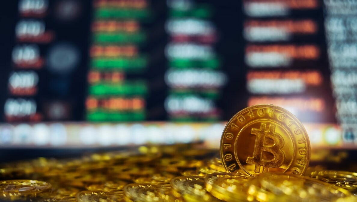 Eine Bitcoin-Münze mit vielen weiteren, die vor einem Bildschirm mit roten und grünen Zahlen liegen.