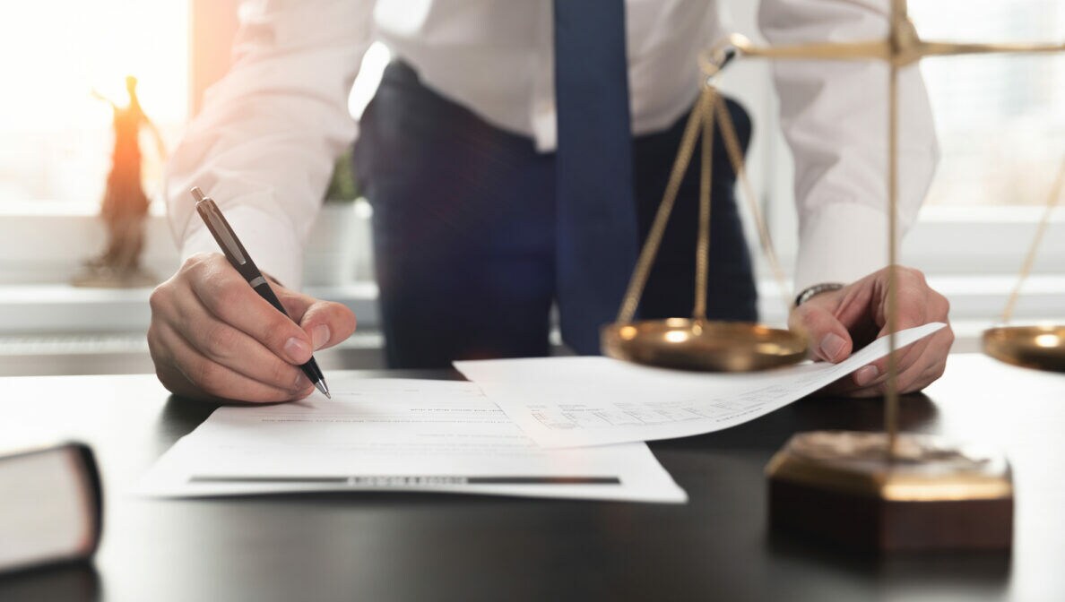 Eine Person im Anzug unterschreibt Dokumente; im Vordergrund steht eine Waage auf dem Tisch