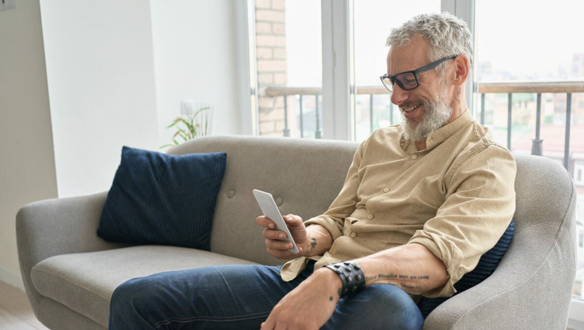 Eine Person mit Brille und grauen Haaren sitzt auf einem Sofa und blickt auf sein Smartphone, das er in seiner Hand hält.