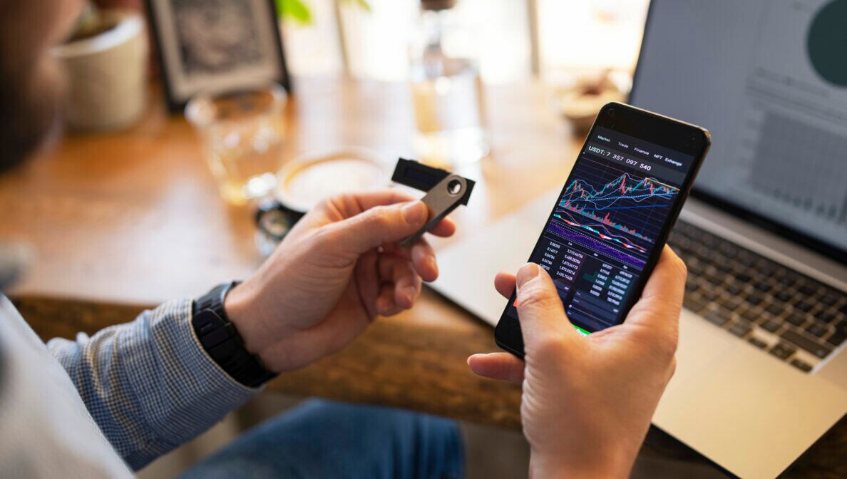 Aufnahme über die Schulter einer Person, die ein Smartphone und einen USB-Stick in den Händen hält. Im Hintergrund steht ein Laptop.