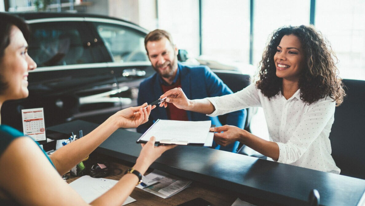 Zwei Personen sitzen im Autohaus am Schreibtisch einer anderen Person gegenüber und überreichen sich gegenseitig Dokumente und Autoschlüssel