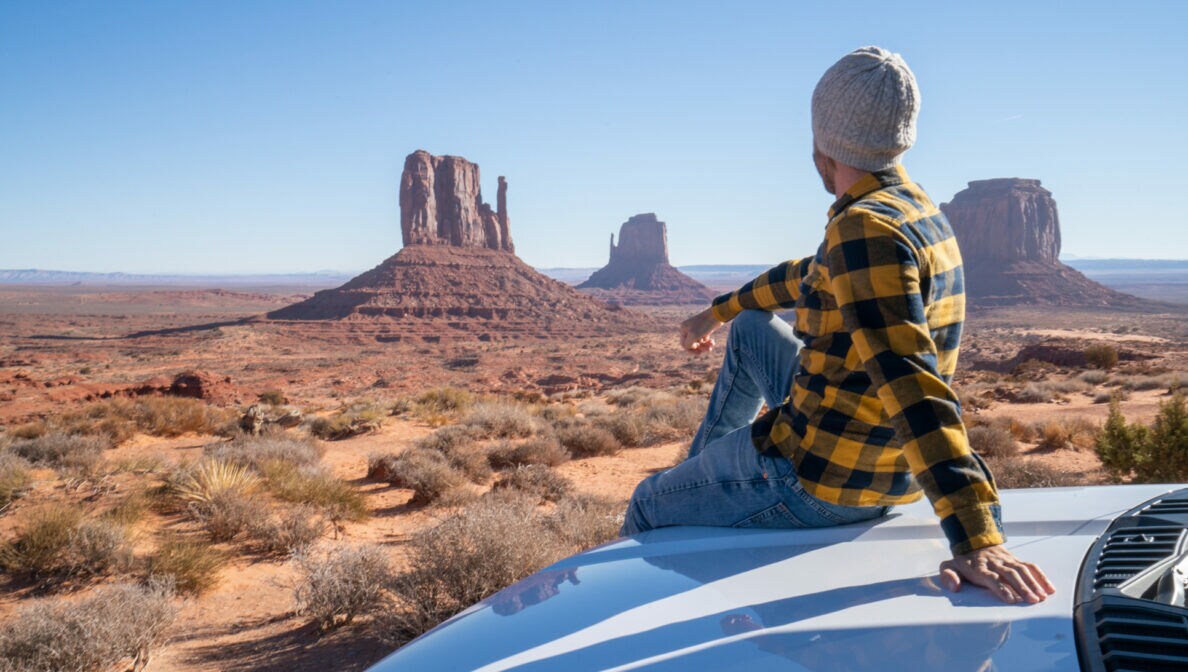 Eine Person sitzt auf der Motorhaube eines Autos und blickt in die Weiten eines US-amerikanischen Nationalparks