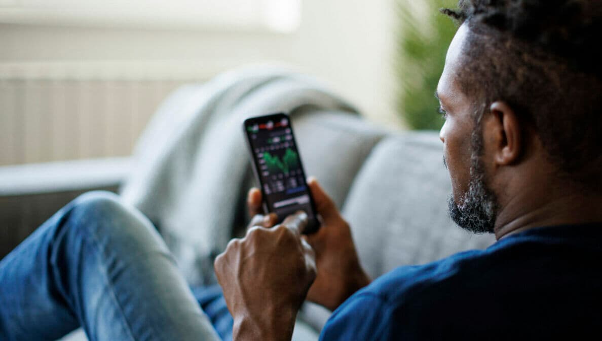 Eine Person sitzt auf einem Sofa und blickt auf sein Smartphone, auf dem Aktienkurse zu sehen sind.