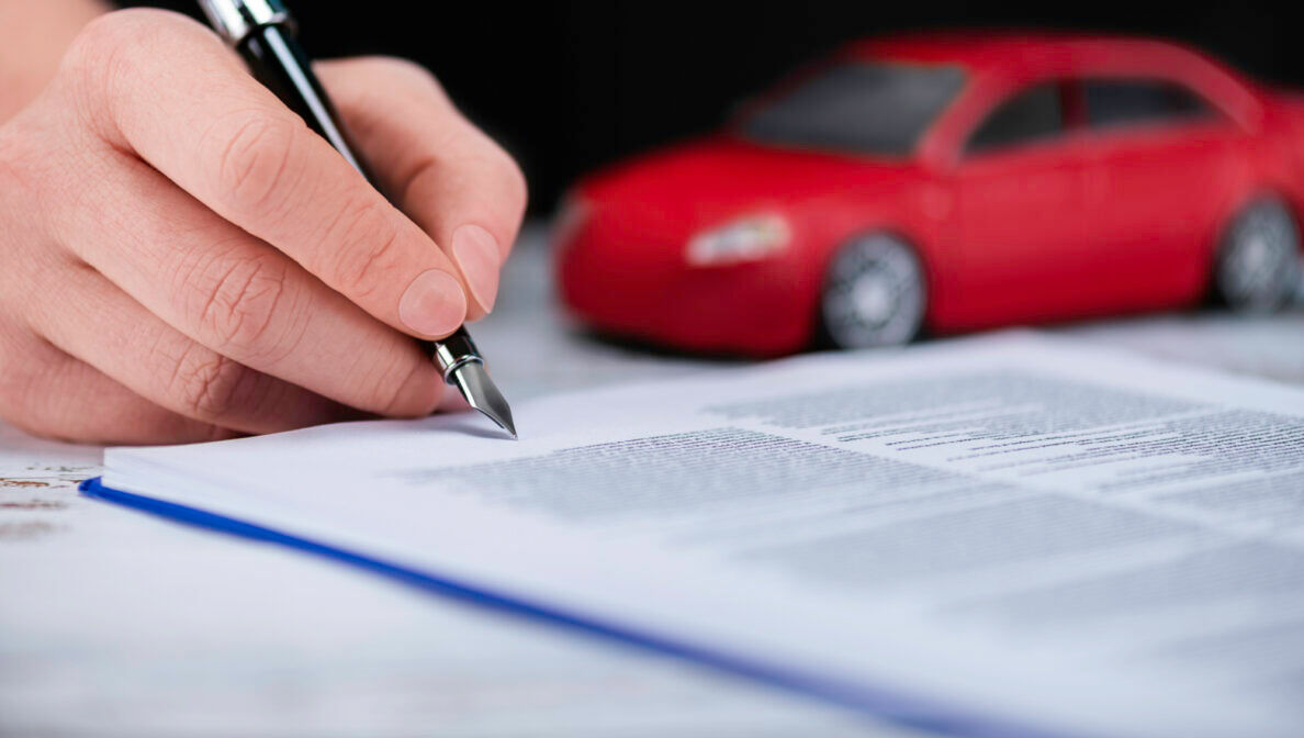 Nahaufnahme einer Hand, die mit einem Füllfederhalter ein Dokument unterschreibt, im Hintergrund steht ein rotes Modellauto.