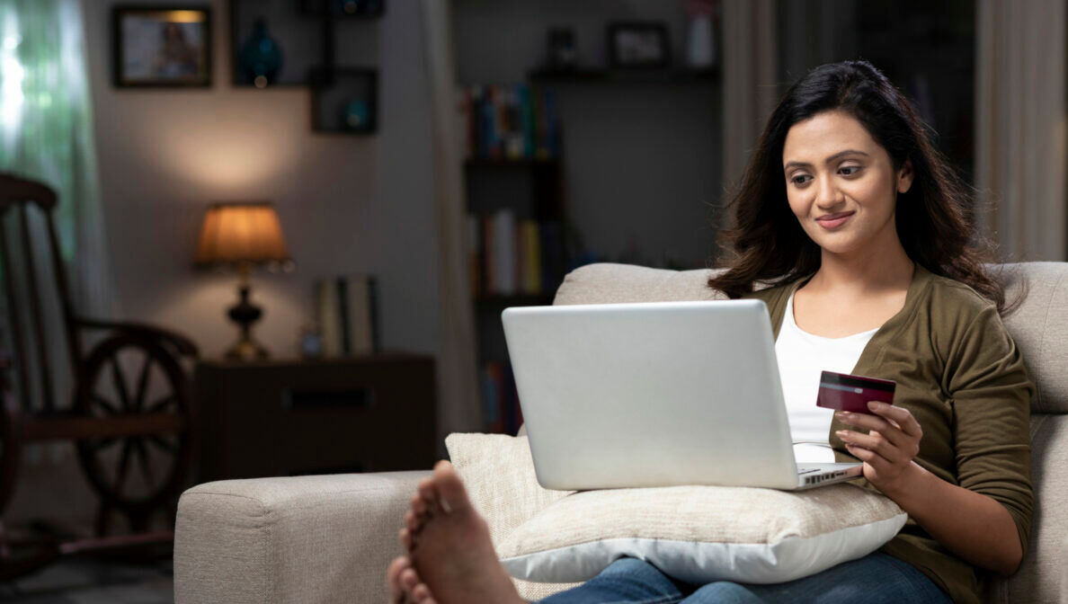Eine Person sitzt mit Laptop auf dem Sofa und hält eine Kreditkarte in der Hand.