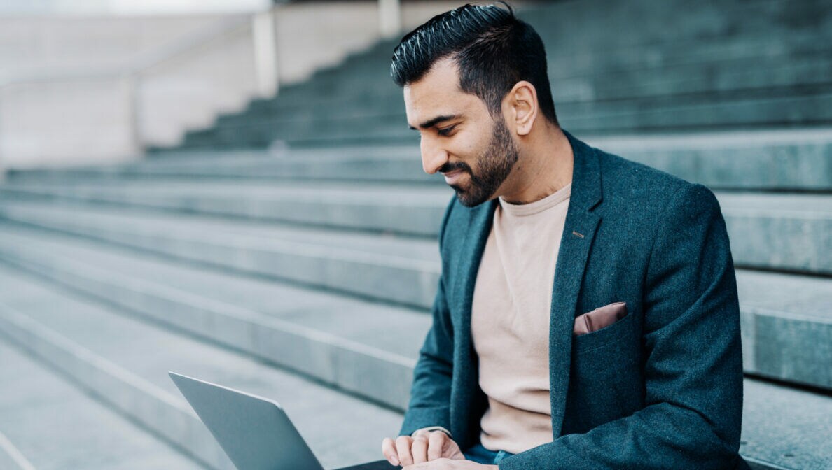Junger, dunkelhaariger Mann mit gepflegtem Bart und Sakko sitzt auf einer Treppenstufe und schaut auf einen aufgeklappten Laptop, der auf seinen Beinen liegt