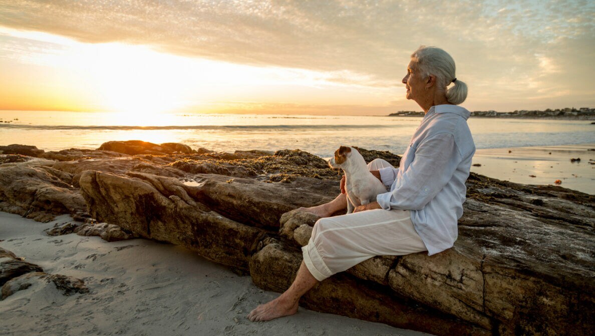 Eine ältere Frau sitzt mit ihrem Hund am Strand und beobachtet den Sonnenuntergang