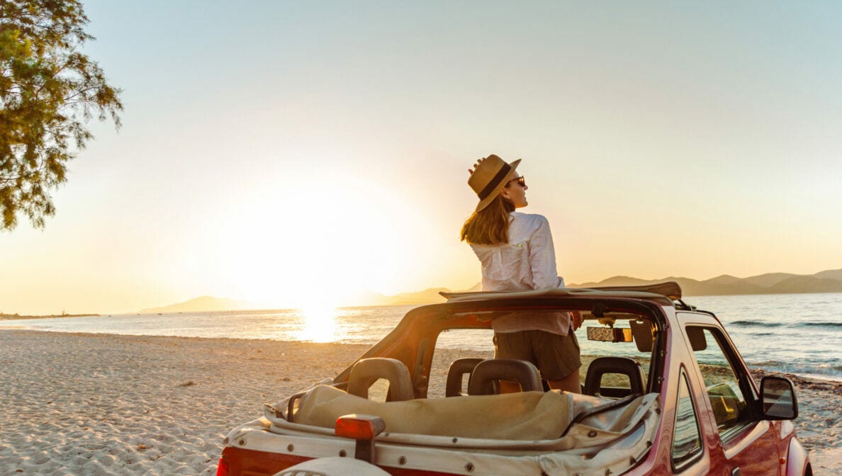 Eine Person in der Rückenansicht, die in einem offenen Geländewagen am Strand steht und den Sonnenuntergang beobachtet.