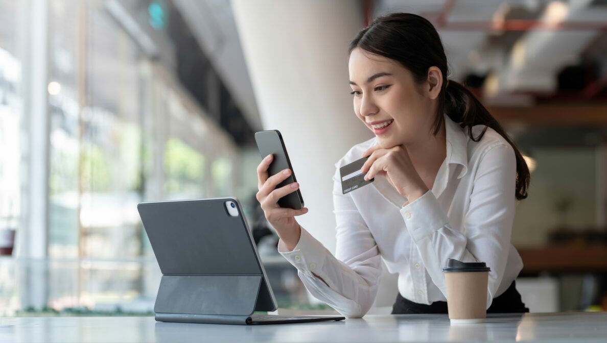 Eine lächelnde Person schaut auf das Smartphone, das sie in der Hand hält. In der anderen Hand hält sie eine Kreditkarte. Vor ihr stehen ein Tablet und ein Becher.