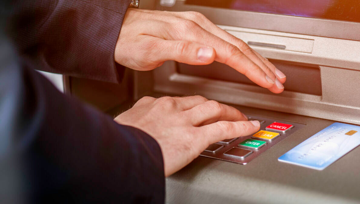 Eine Hand tippt etwas auf einem Bankautomaten ein, die andere Hand liegt schützend darüber.