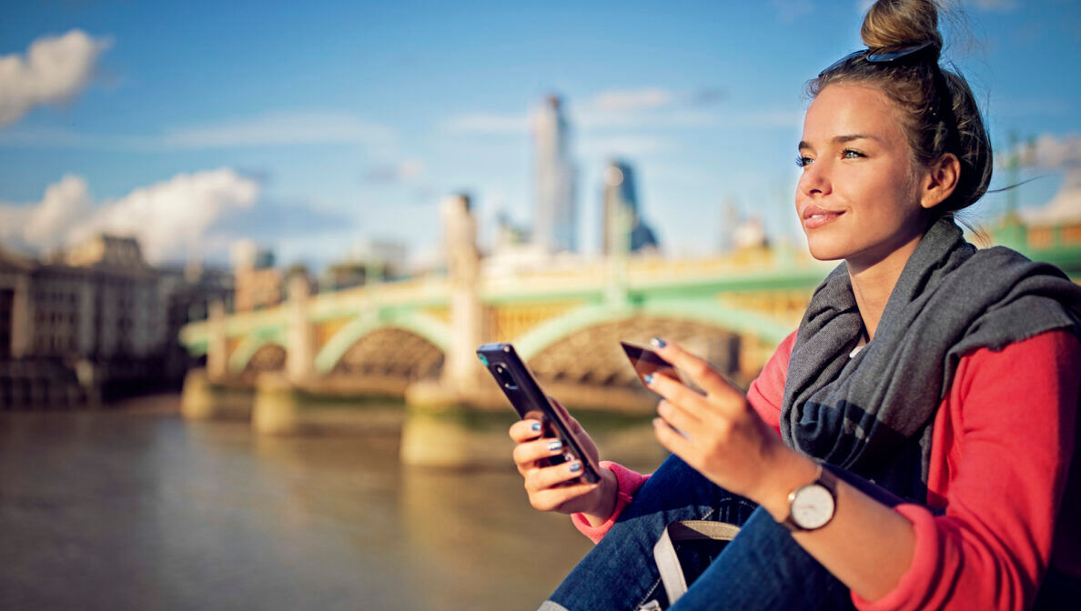 Eine junge Frau mit Haarknoten sitzt in einer Stadt an einem Fluss vor einer Brücke. In der Hand hält sie eine Kreditkarte und ein Handy.