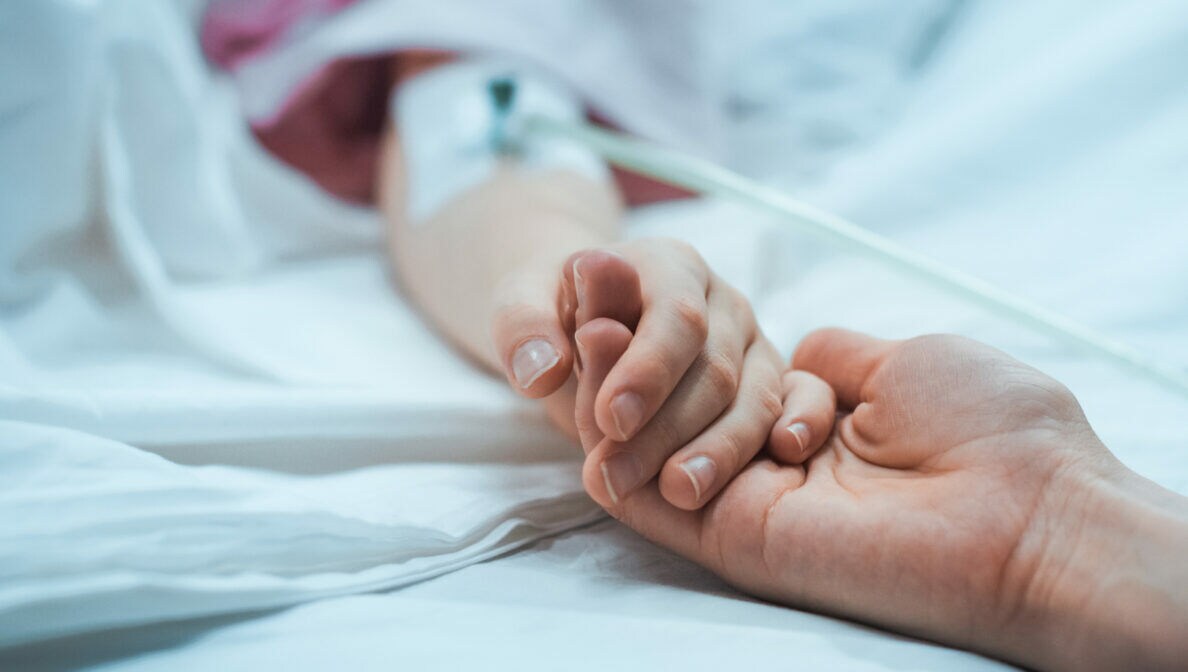Zwei Personen halten sich an den Händen, von denen eine im Krankenbett liegt und eine Infusion bekommt