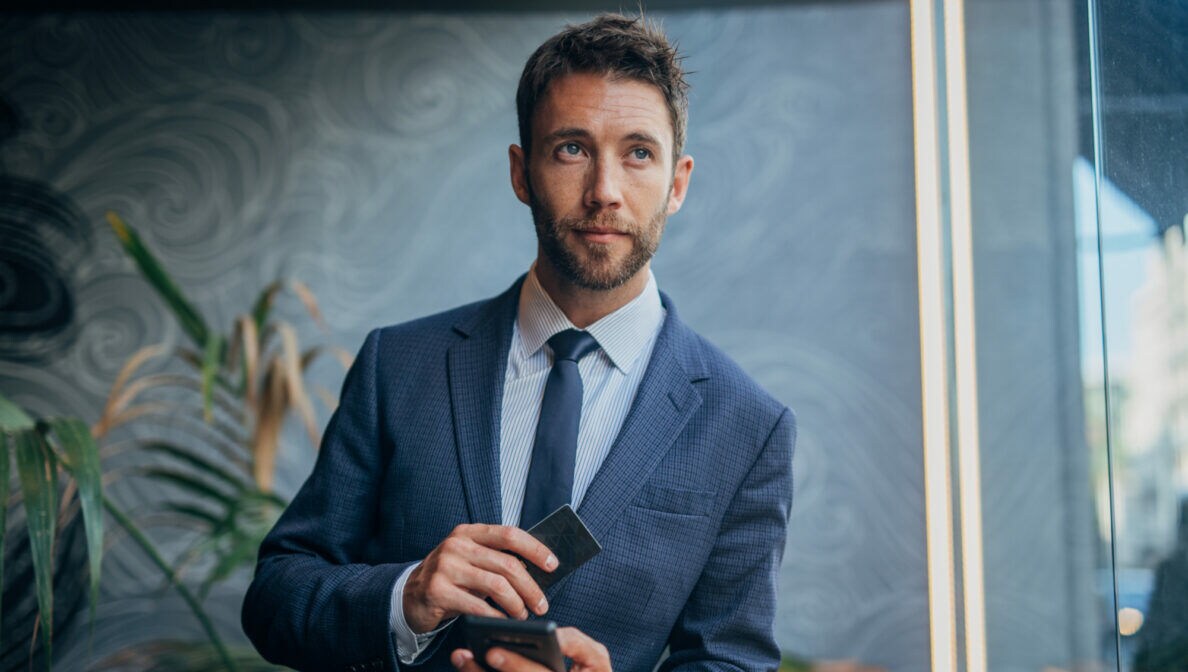 Eine Person in dunkelblauem Anzug mit Krawatte lehnt auf einem Tresen, auf dem eine Espressotasse steht. Sie hält ein Handy und eine Bezahlkarte in Händen.