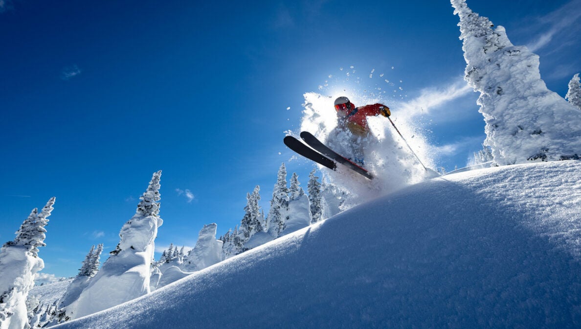 Eine Person springt mit seinen Skiern über einen kleinen Schneehügel, hinter ihm scheint die Sonne.