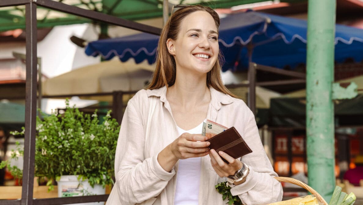 Eine Frau mit einem geflochtenen Einkaufskorb am Arm, die zwei Euroscheine aus einem Geldbeutel entnimmt.