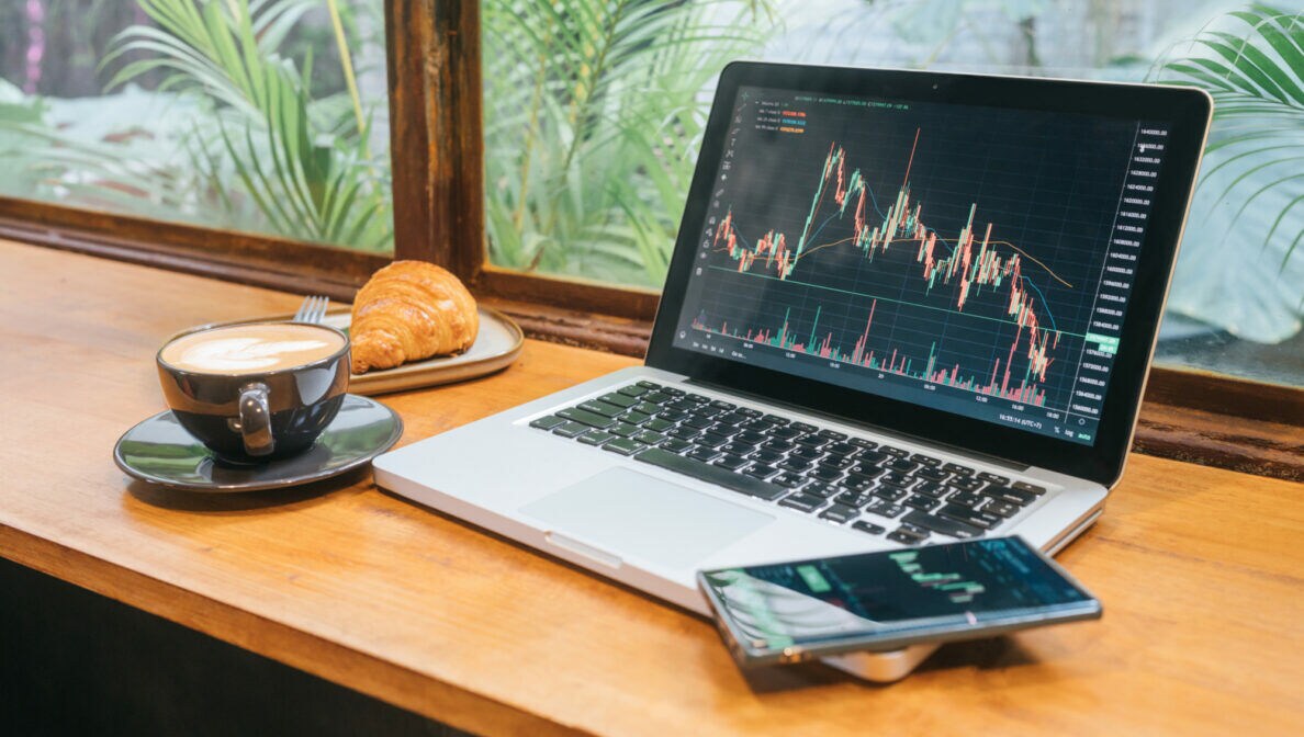 Auf einem Tisch am Fenster steht ein aufgeklappter Laptop und Smartphone. Auf den Bildschirmen sind Kurswerte abgebildet. Daneben stehen eine Tasse Kaffee und ein Teller mit einem Croissant.