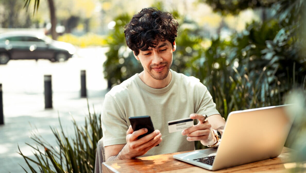 Ein junger Mann sitzt an einem Tisch, vor ihm steht ein aufgeklappter Laptop. In einer Hand hält er ein Smartphone, in der anderen eine Kreditkarte.