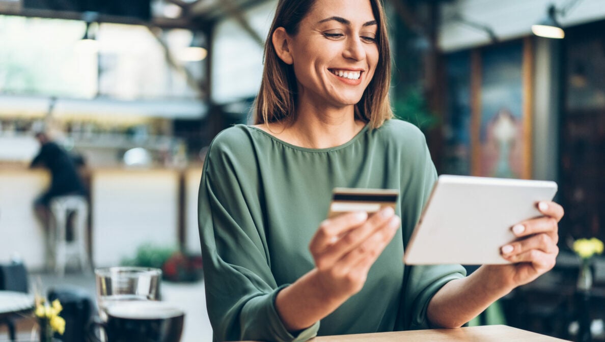 Eine Frau sitzt in einem Café und hält lächelnd eine Karte und ein Tablet vor sich