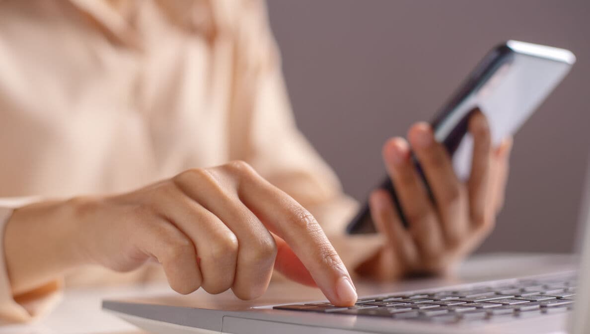Eine Frau hält ein Handy in der linken Hand und drückt mit der rechten Hand eine Taste auf ihrem Laptop