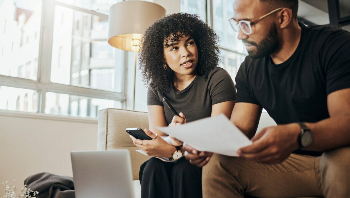 Zwei farbige Menschen sitzen nebeneinander auf einem Sofa. Der Mann hält ein Blatt, die Frau ein Handy in der Hand. Vor ihnen auf dem Tisch steht ein aufgeklappter Laptop
