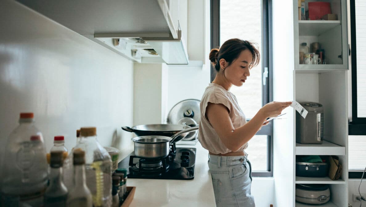 Eine junge Frau steht in einer Küche. Sie sieht sich einen Zettel an, den sie in der Hand hält.