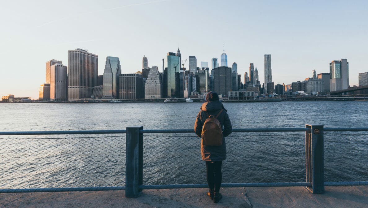 Eine dick angezogene Frau mit Rucksack steht vor einem Geländer. Sie betrachtet einen Fluss und die Skyline von Manhattan