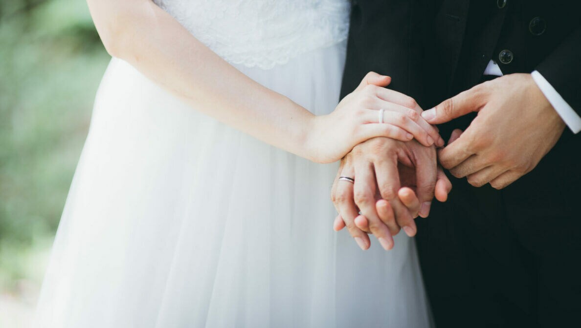 Nahaufnahme von Händen mit Eheringen. Sie gehören einer Frau im Brautkleid sowie einem Mann im Anzug.