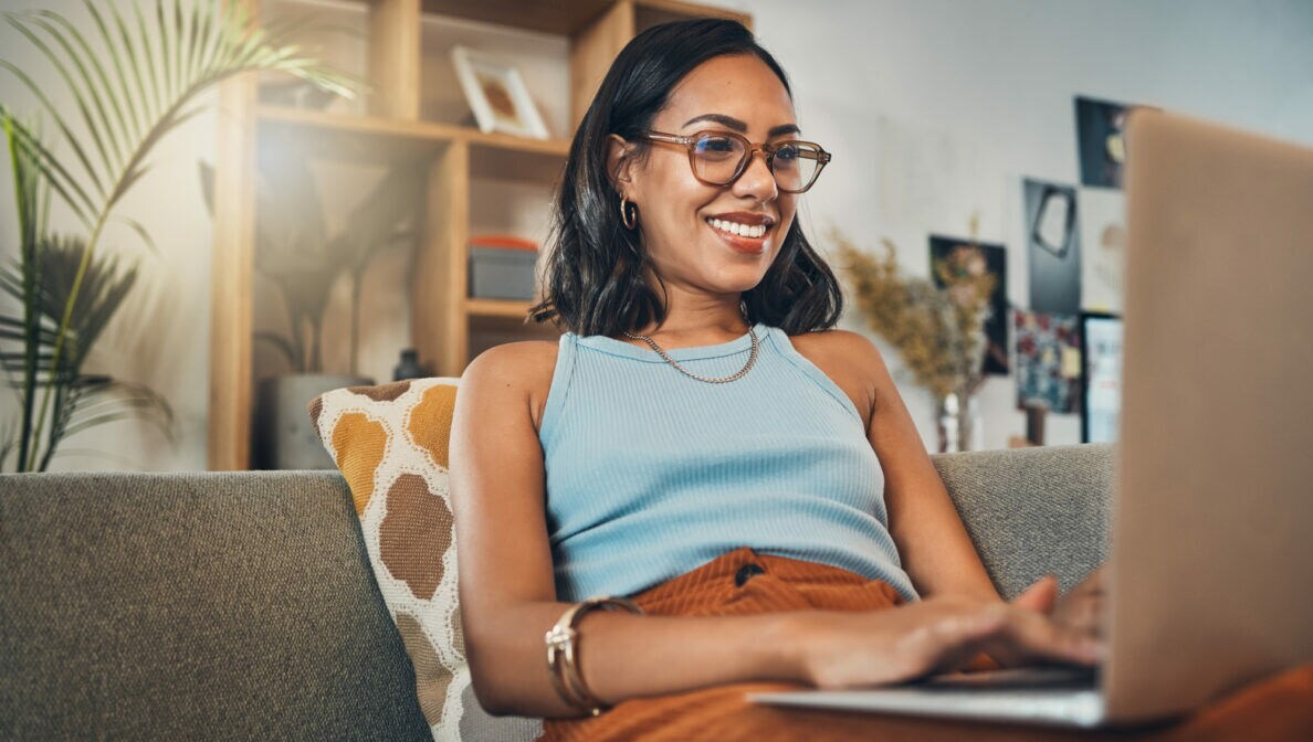 Eine Frau sitzt auf dem Sofa und hat einen aufgeklappten Laptop auf dem Schoß.