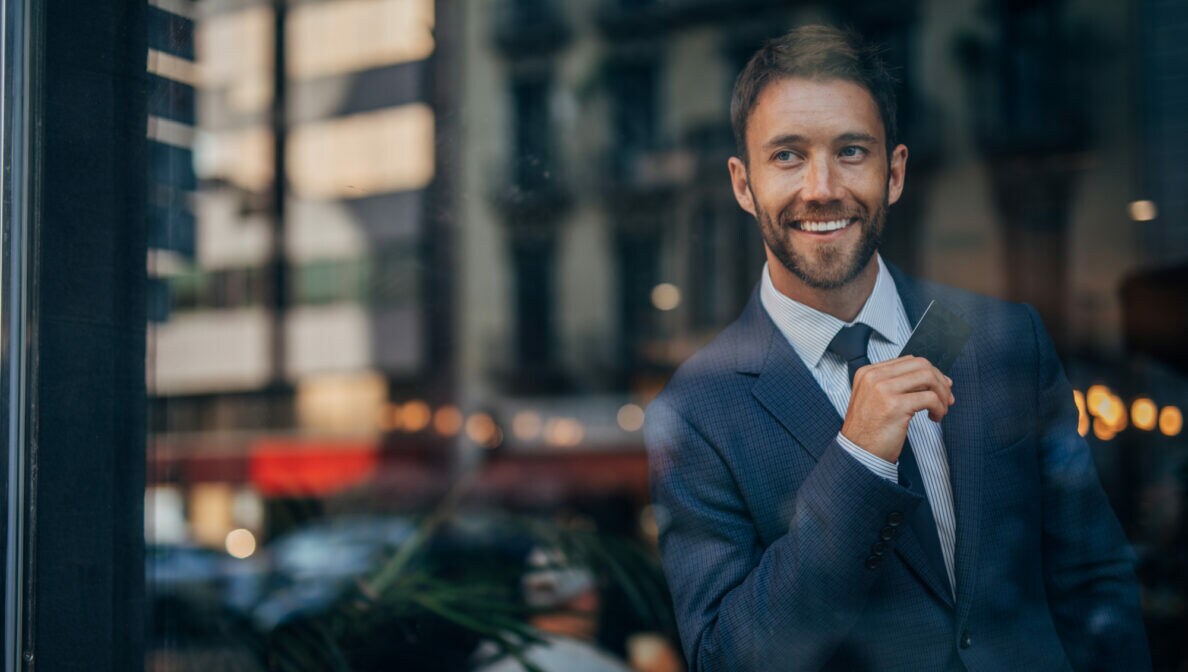 Eine Person im Anzug steht in einem Café und blick aus dem Fenster, während er eine Kreditkarte in der Hand hält.