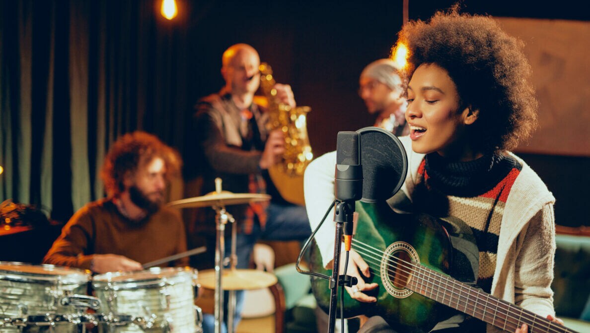 Eine Person sitzt in einem kleinen Club mit ihrer Gitarre auf einem Hocker und singt in ein Mikrofon.