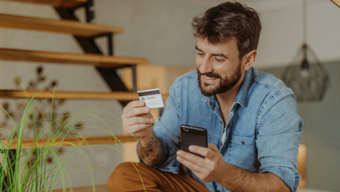 Ein Mann sitzt lächelnd auf den Stufen einer Treppe. In der einen Hand hält er eine Kreditkarte, auf die er schaut. In der anderen Hand hält er ein Smartphone