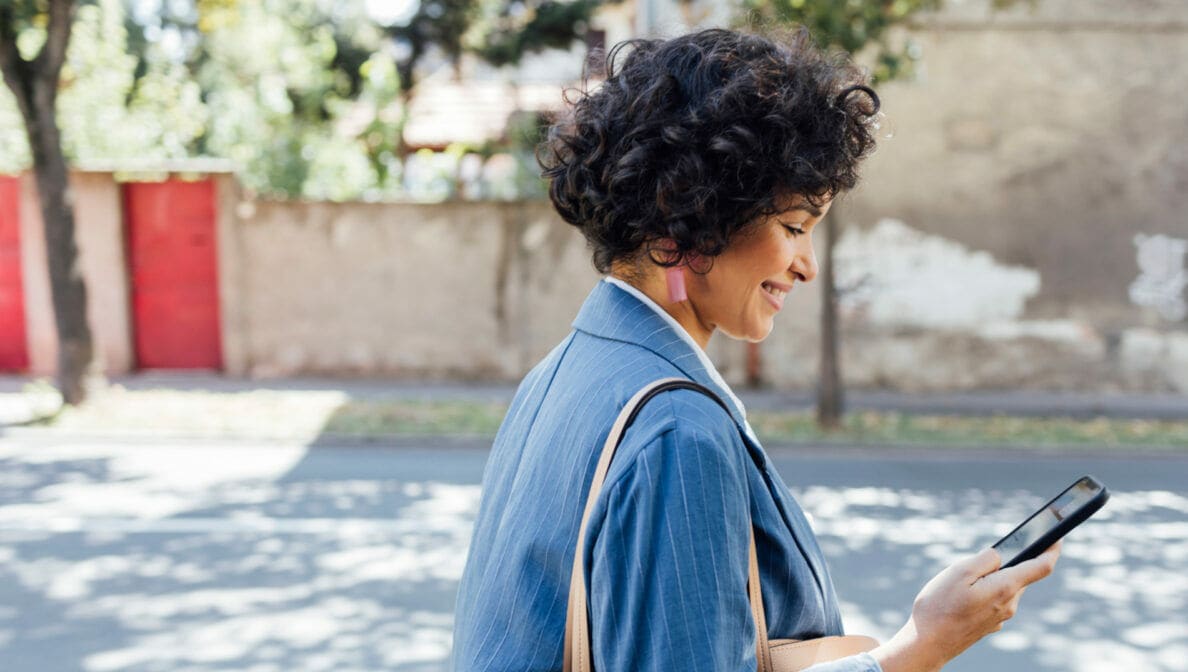 Eine Frau geht an einer Straße spazieren und schaut dabei lächeld auf ihr Smartphone