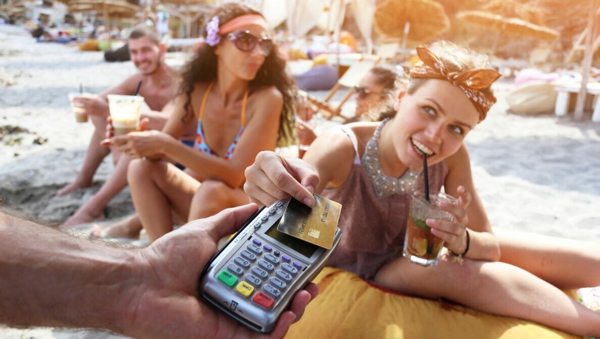 Zwei fröhliche, junge Personen sitzen am Strand und halten Getränke in der Hand. Eine der Personen legt eine Kreditkarte auf ein Kartenlesegerät