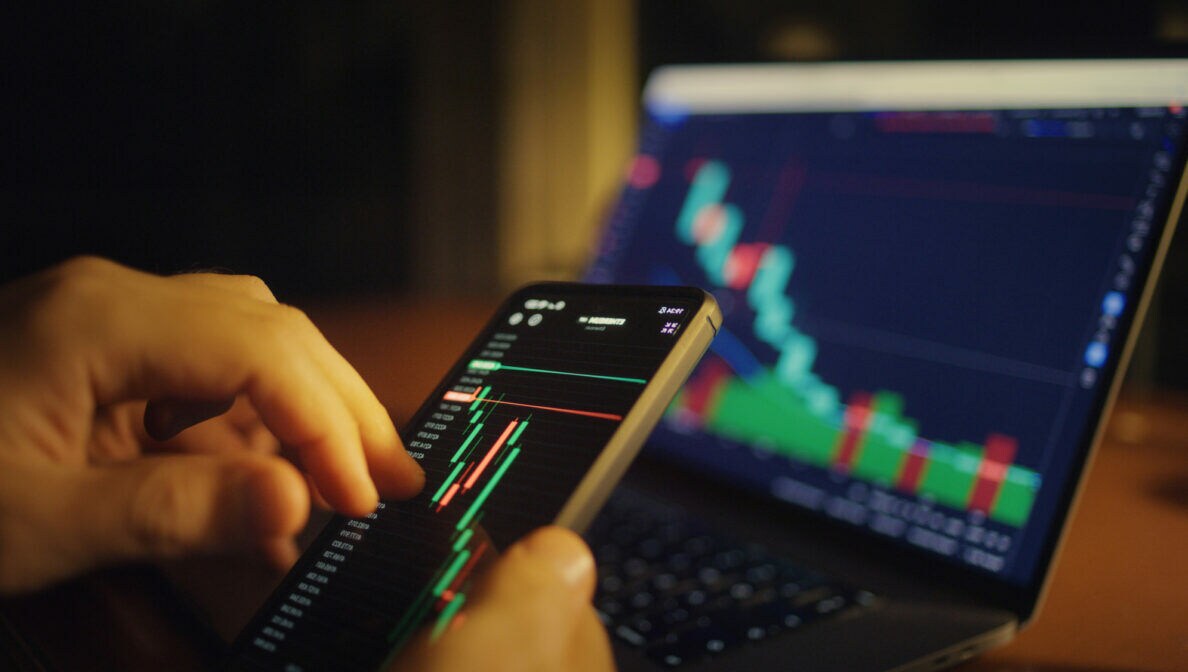 Zwei Hände halten ein Handy vor einen Laptop. Auf beiden Geräten sind Börsenergebnisse zu sehen