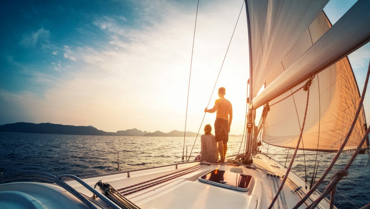 Zwei Silhouetten sitzen und stehen auf einem Segelboot, während das Boot in den Sonnenuntergang segelt