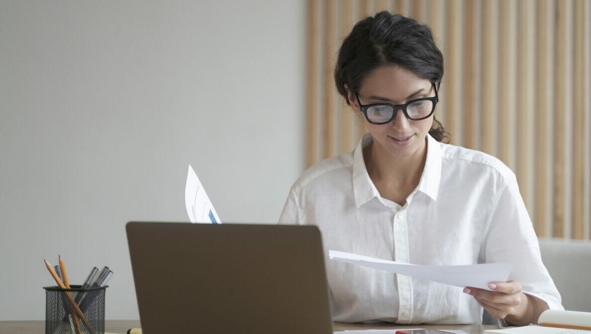 Eine Person sitzt am Schreibtisch mit einem Laptop und hält Zettel in der Hand