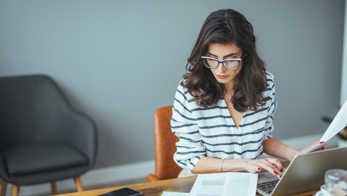 Eine Person sitzt am Schreibtisch vor einem geöffneten Laptop, vor ihr liegen Dokumente und Geldscheine.