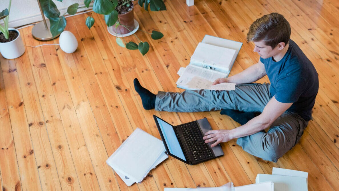 Eine Person sitzt auf dem Fußboden mit Aktenordnern und einem Laptop und wird von oben fotografiert.