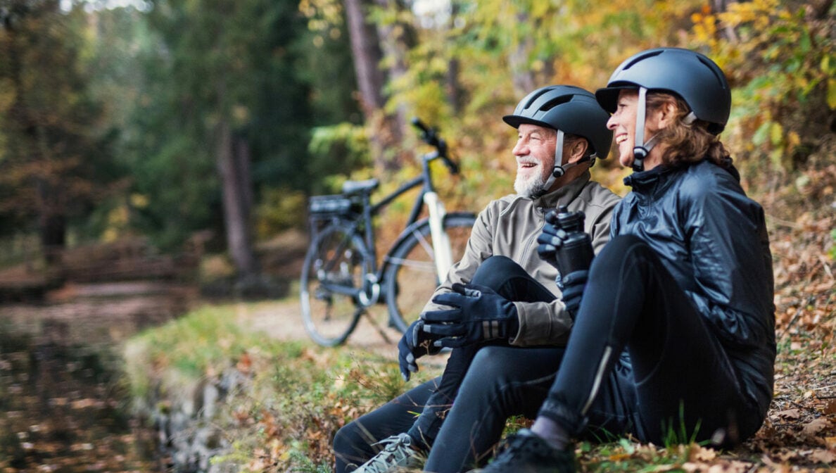 Zwei ältere Personen sitzen nebeneinander an einem Waldweg auf dem Boden. Sie tragen Fahrradhelme und im Hintergrund steht ein Fahrrad.