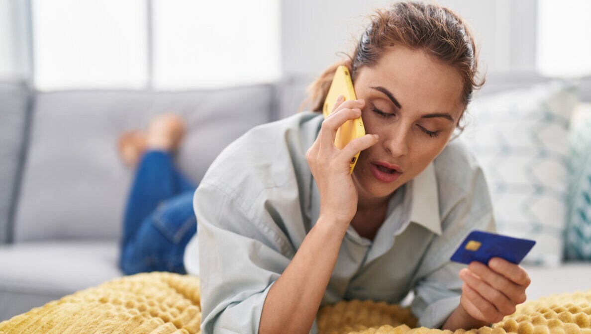 Eine Person liegt bäuchlings und telefonierend auf dem Sofa und hält eine Kreditkarte in der Hand.