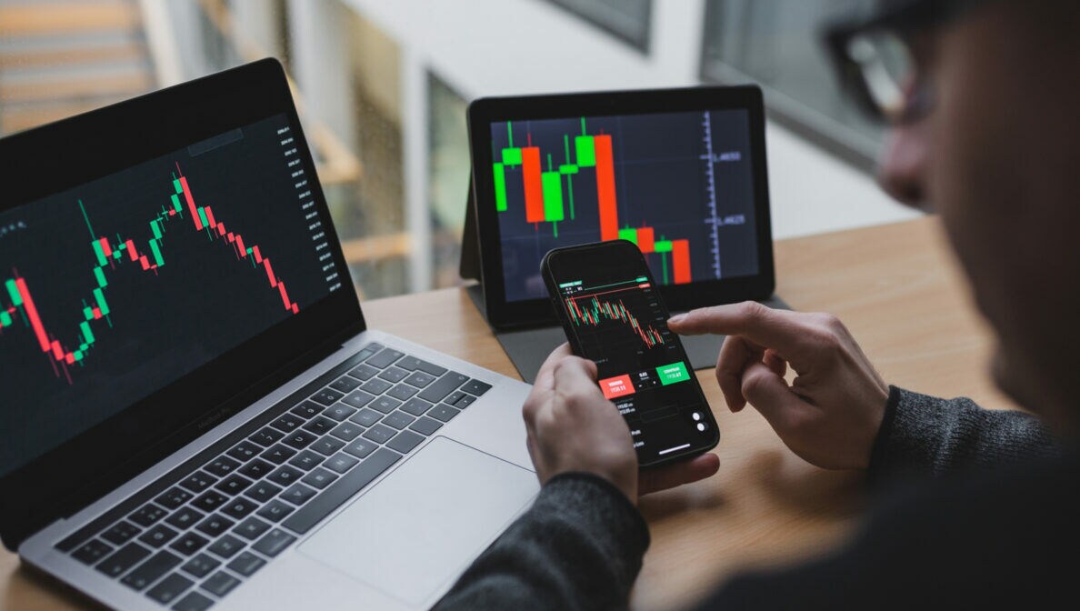 Eine Person sitzt am Schreibtisch, auf dem ein Laptop und Tablet mit Börsenkursen stehen. In der Hand hält er ein Smartphone mit den gleichen Darstellungen.