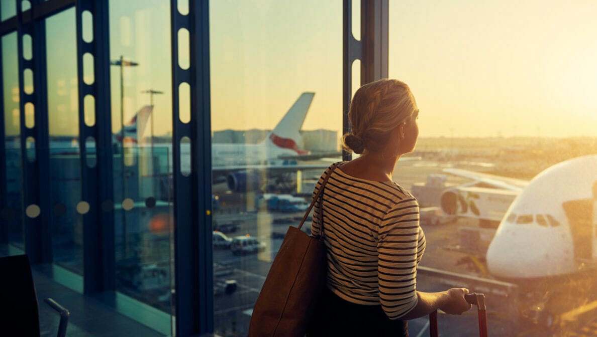 Eine Person steht im Flughafen und blickt aus dem Fenster, vor dem mehrere Flugzeuge stehen.