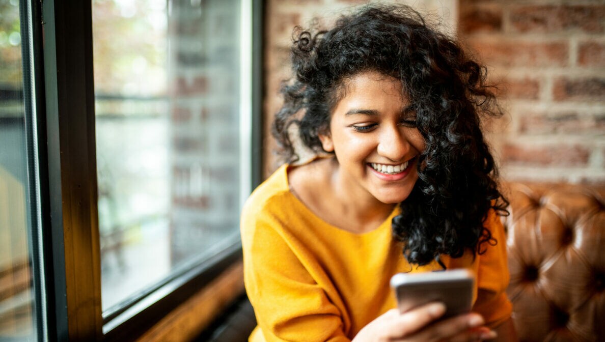 Eine Person sitzt auf einem Ledersofa und blickt lächelnd auf ihr Smartphone.