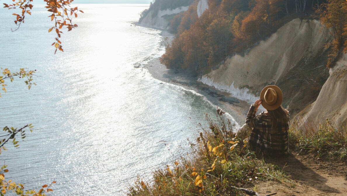Eine Person mit großem Hut sitzt an einer Klippe mit Blick Richtung Meer.