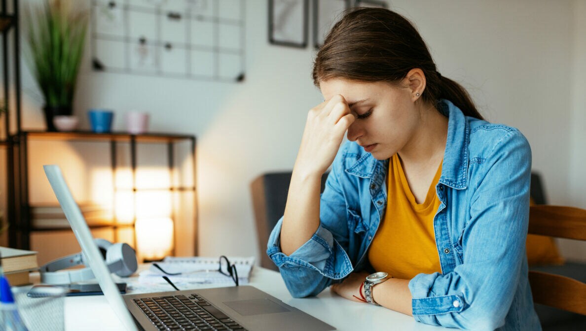 Eine Frau sitzt am Tisch vor einem Laptop und stützt den Kopf mit einer Hand.