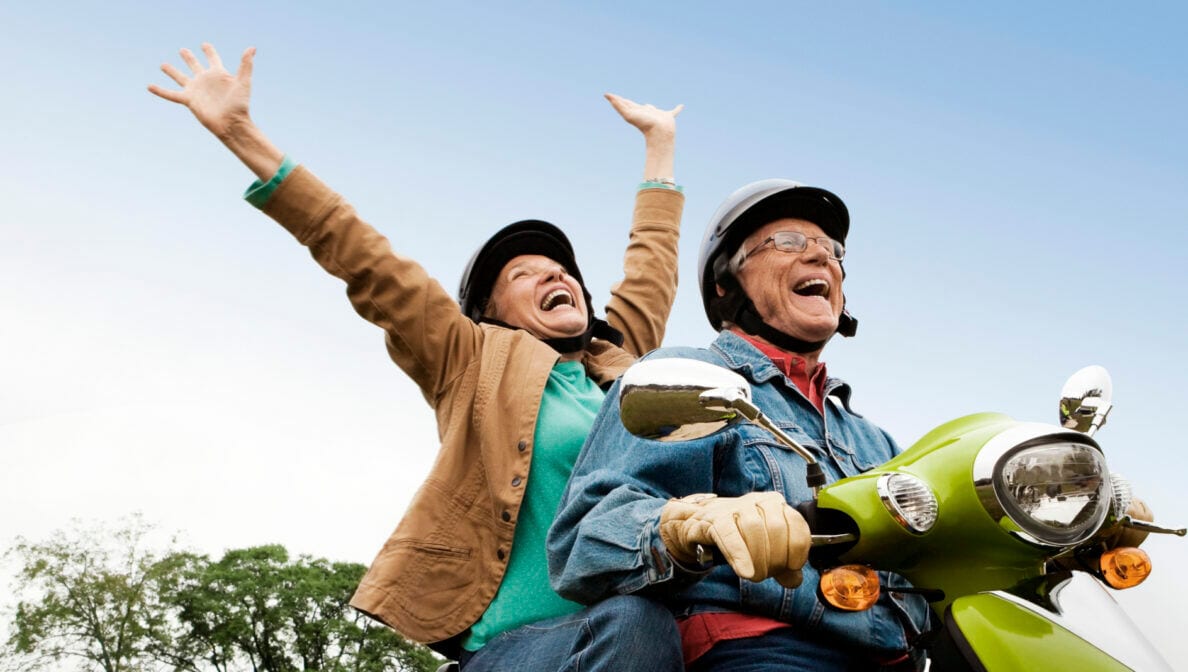 Ein älterer Mann und eine ältere Frau sitzen lachend auf einem grünen Moped. Die Frau streckt beide Arme in die Höhe.
