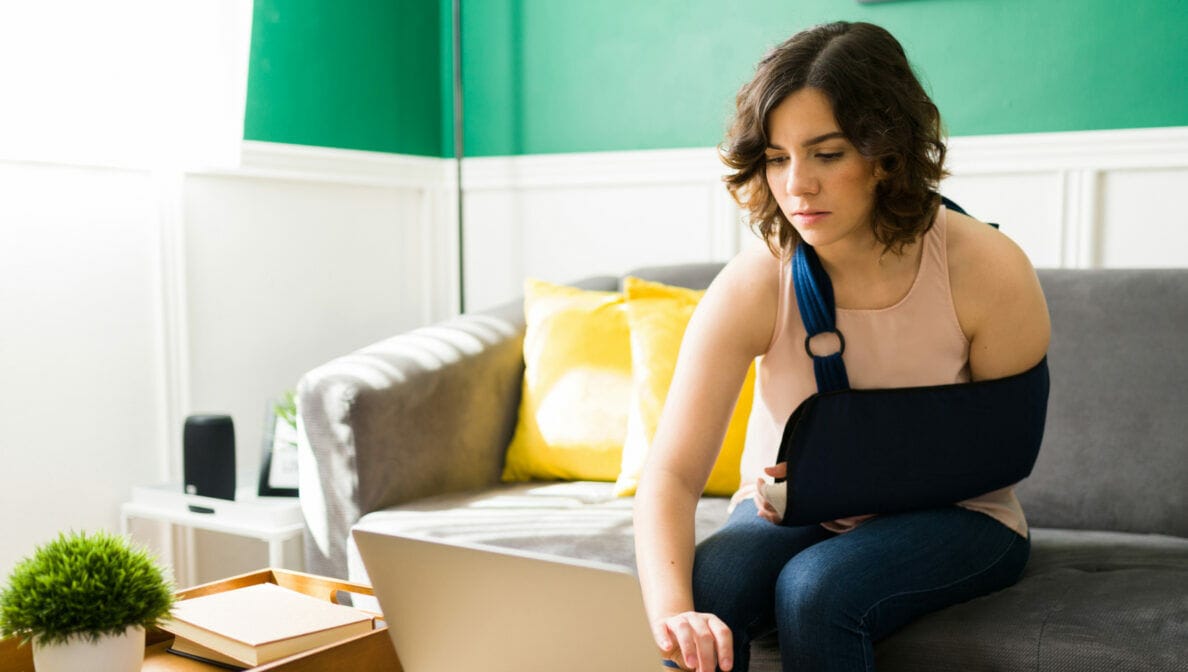 Eine Frau mit ruhiggestelltem Arm sitzt auf einem Sofa und tippt mit der freien Hand auf ihrem Laptop.